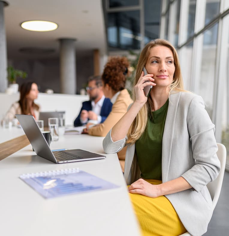 Blond entrepreneur discussing over phone call while sitting in board room with professional teammates in the background Blond entrepreneur discussing over phone call while sitting in board room with professional teammates in the background