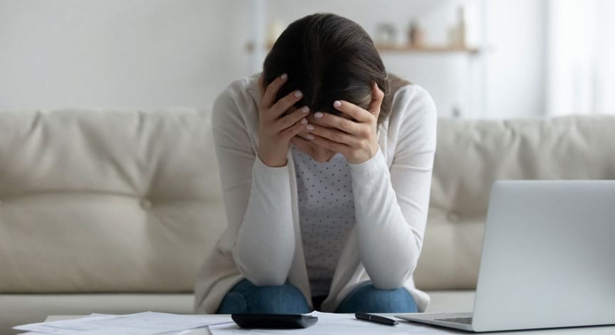 woman-sitting-on-a-couch-holding-her-head-with-paperwork-and-a-laptop-in-front-of-her
