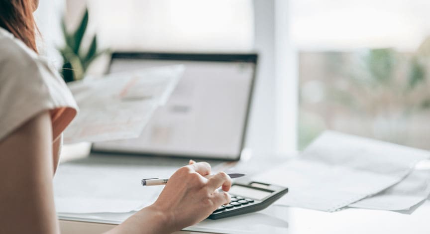 woman-writing-notes-at-a-desk-beside-a-laptop-and-paperwork