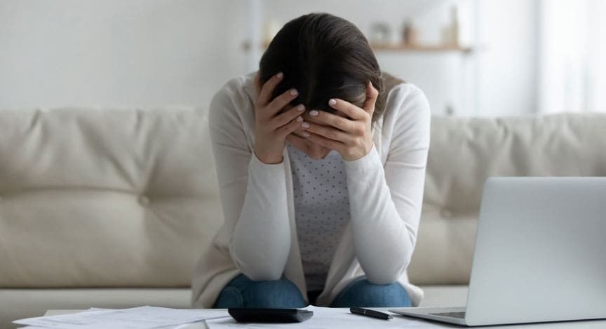 woman-sitting-on-a-couch-holding-her-head-with-paperwork-and-a-laptop-in-front-of-her