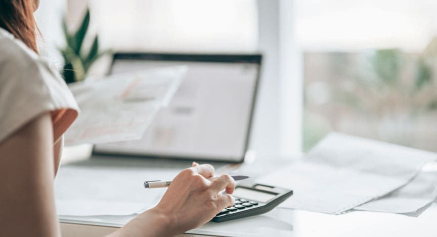woman-writing-notes-at-a-desk-beside-a-laptop-and-paperwork